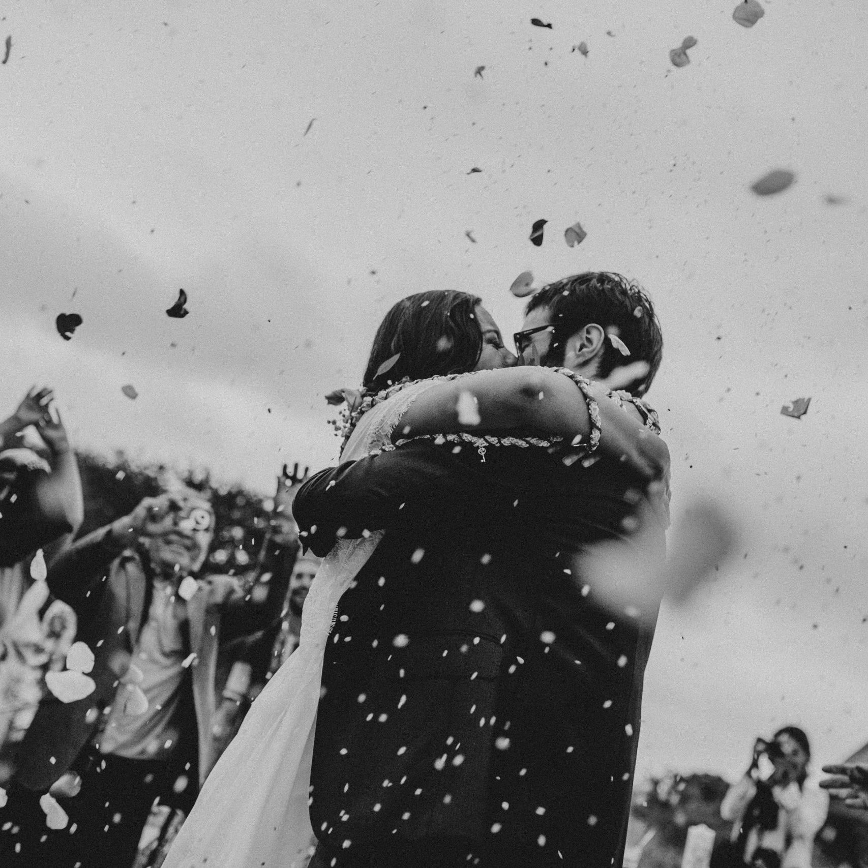 Home Bride and groom embrace in a shower of flower petals during their outdoor wedding ceremony.